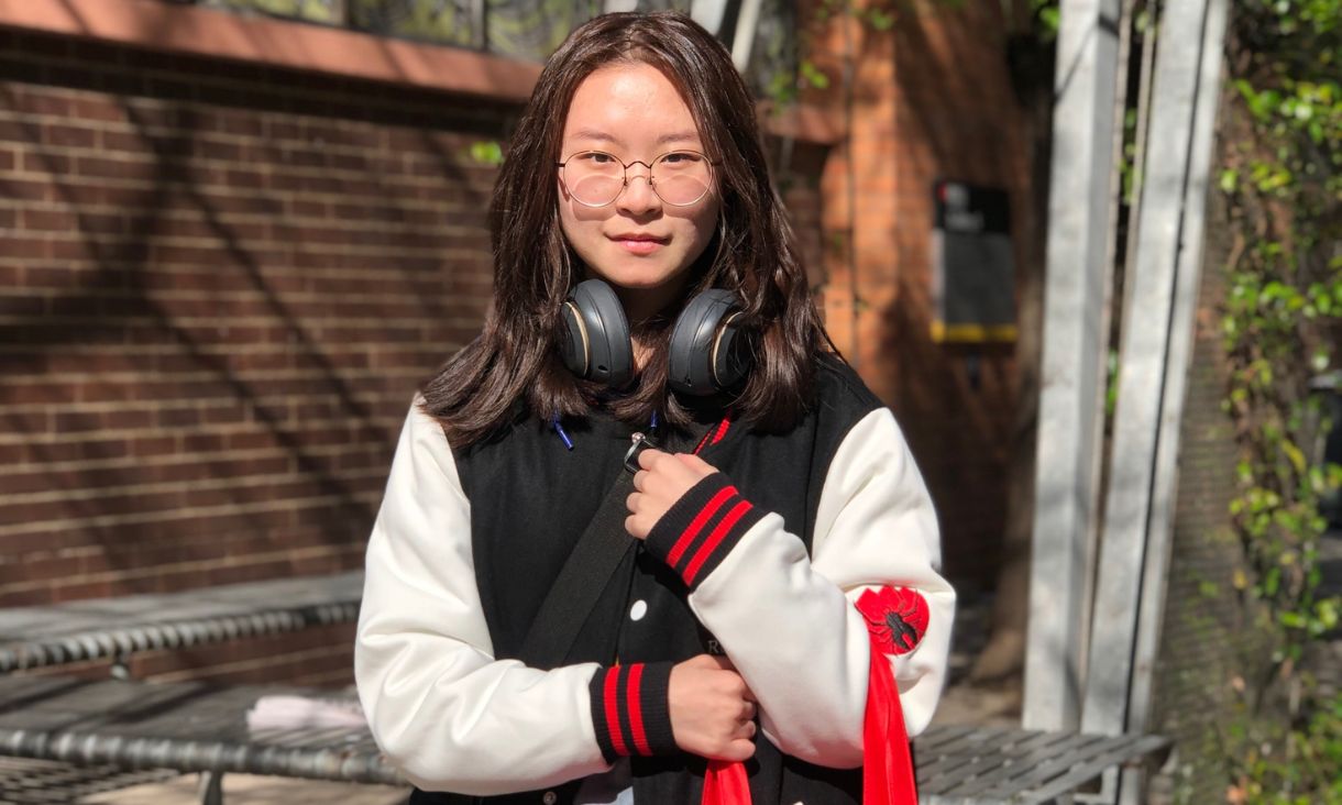 Young Asian female student Xi wearing a varsity RMIT jumper and glasses standing outside holding an RMIT tote bag with gifts.