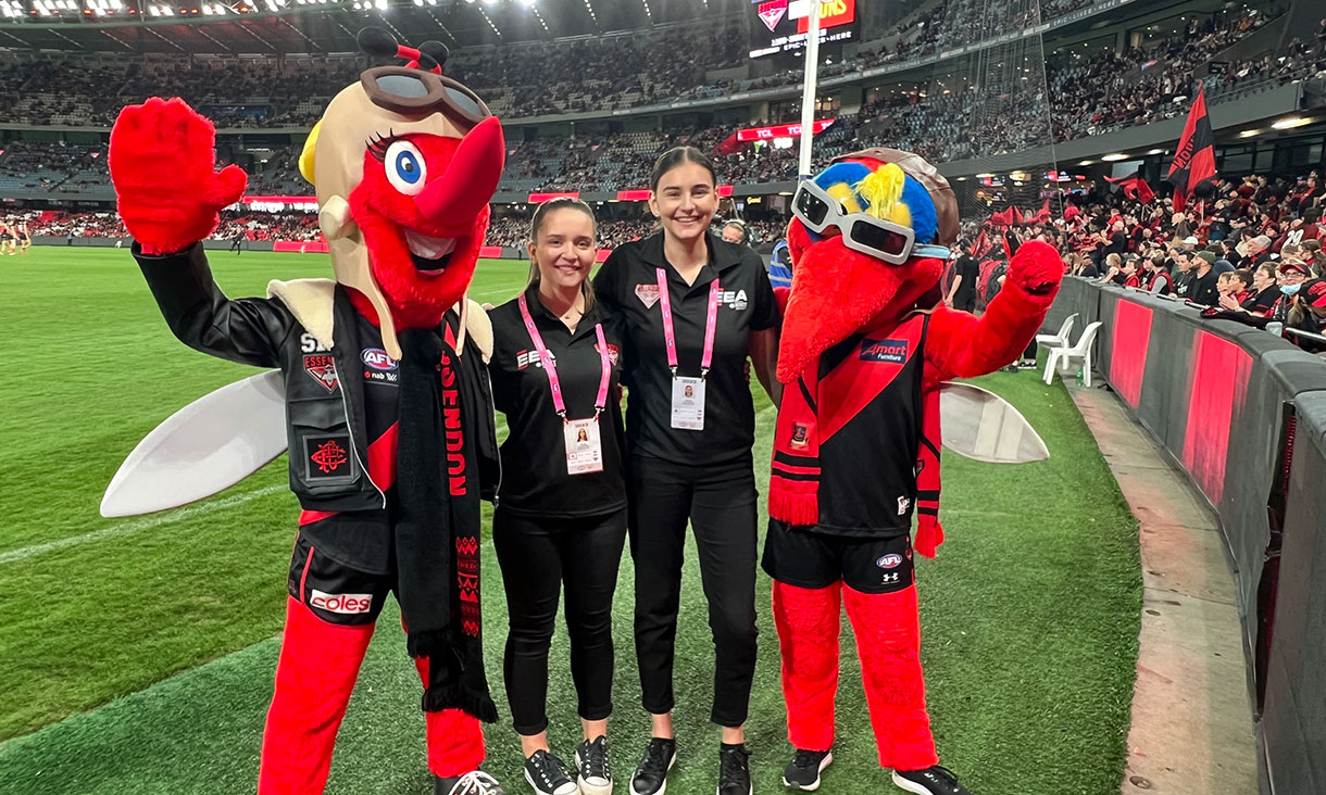 Students standing with Essendon mascots on the grounds of a stadium