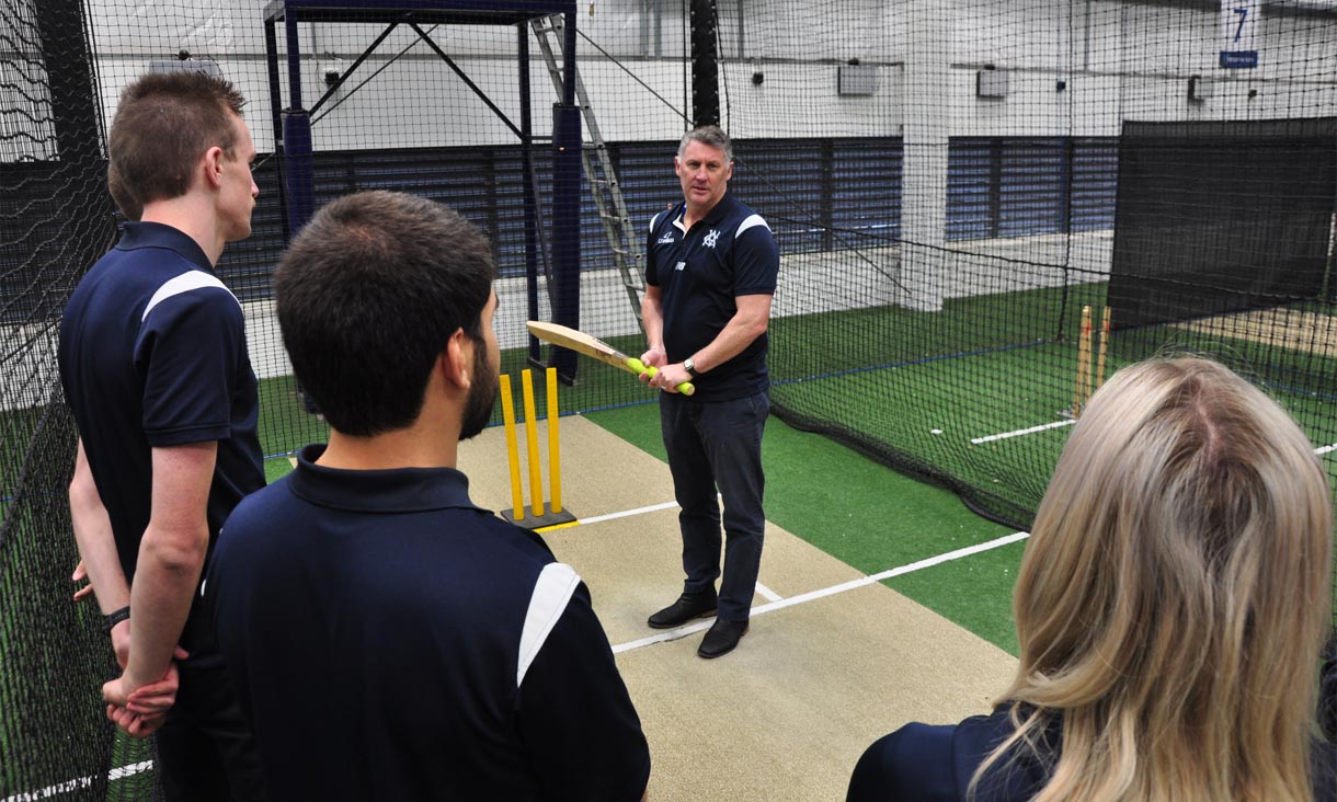 A Cricket Victoria coach wielding a cricket bat on at the crease, inside indoor nets with 3 students watching on