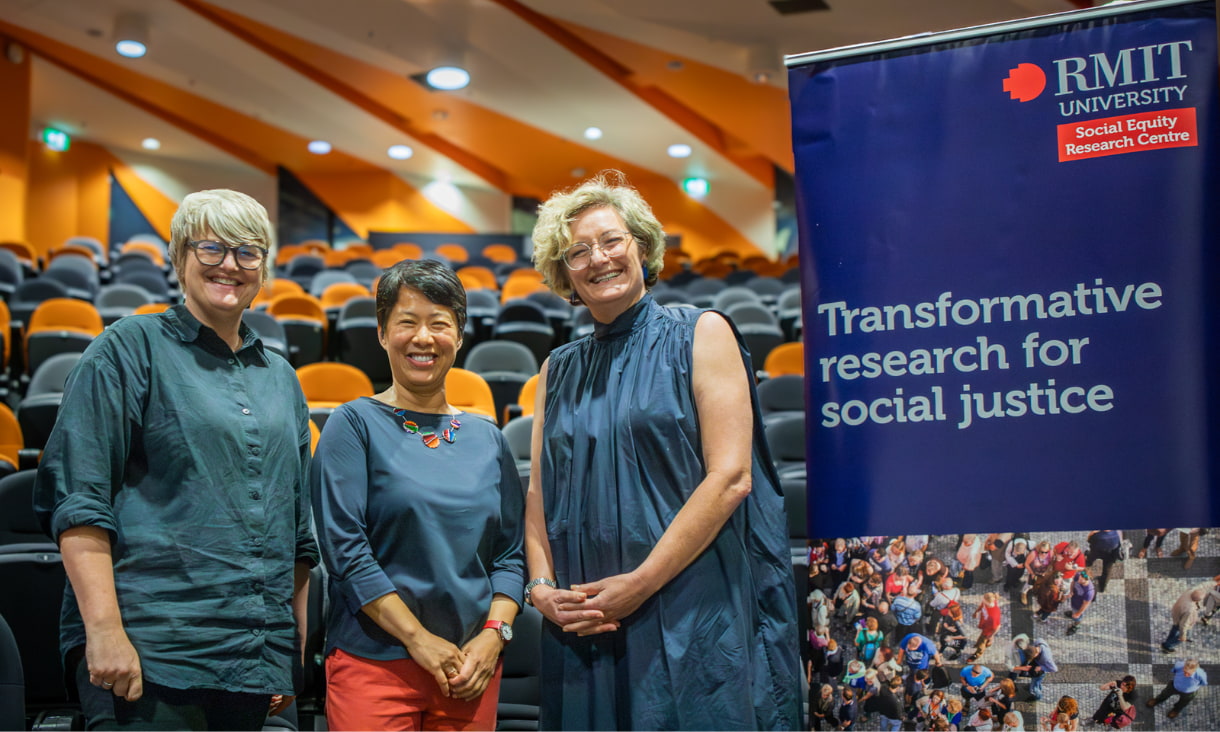 3 women stand in front of an auditorium next to a banner reading 'Transformative research for social justice'.