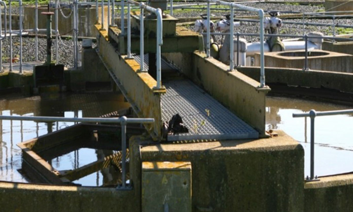 A wastewater treatment facility with a metal walkway over a circular tank of water. Railings line the walkways, and pipes are visible in the background under a sunny sky.