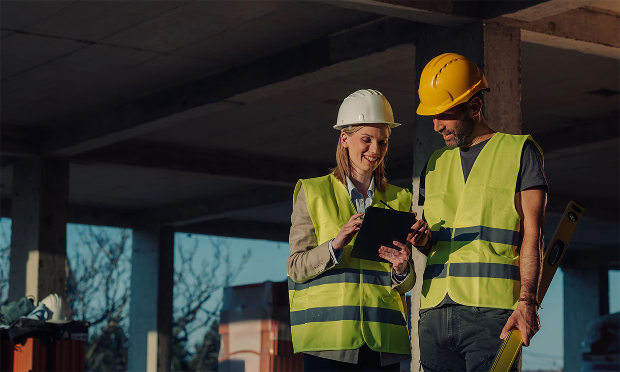 Two engineers wearing safety vests and hard hats reviewing a tablet at a building site.