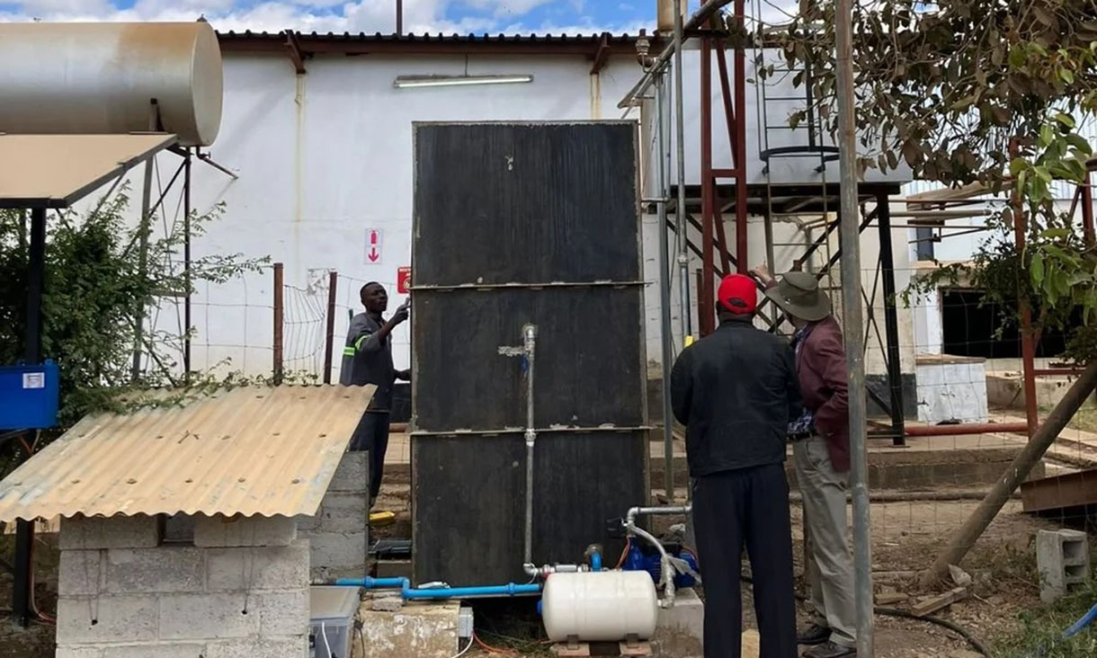 Several individuals are observing and interacting with industrial equipment outdoors. The setup includes pipes and panels, with a building in the background. A large water tank is visible, along with foliage surrounding the area.