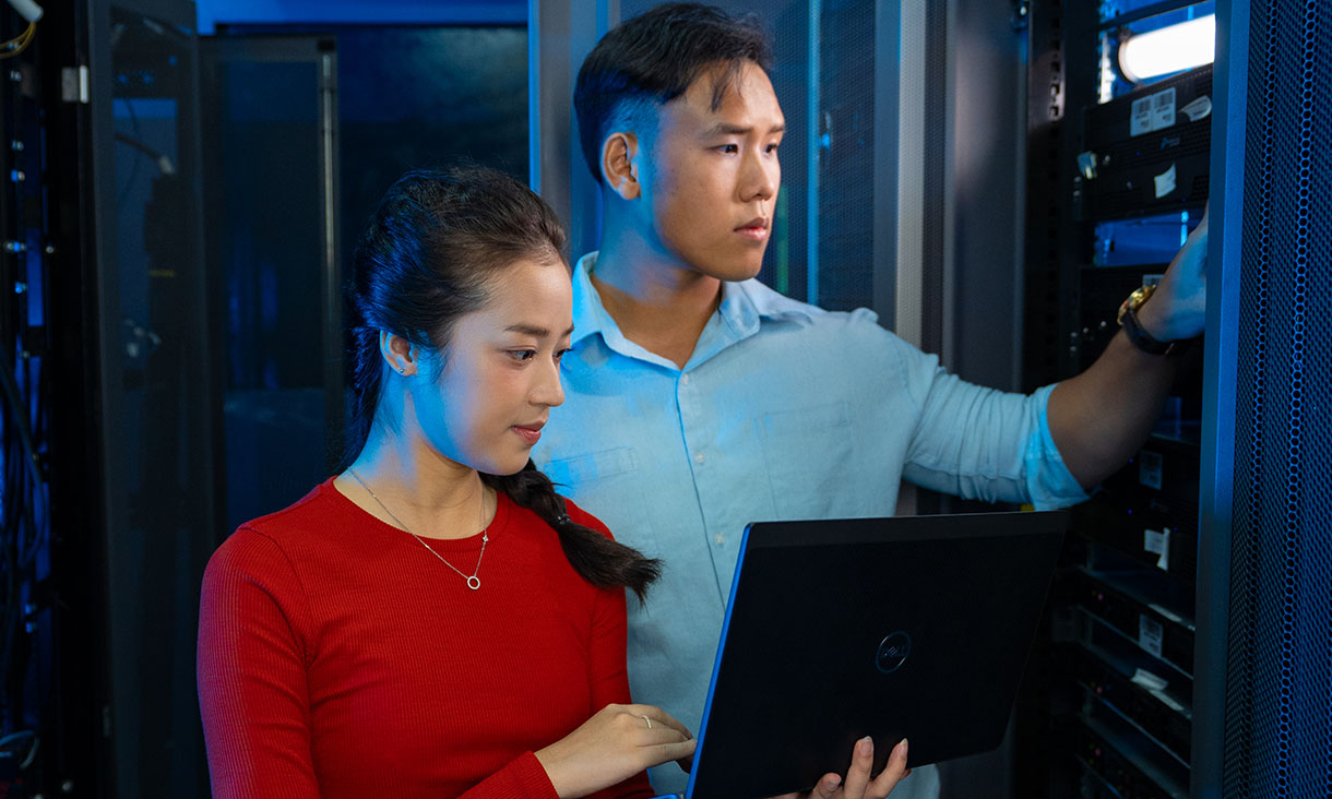 Two people in a server room working with a laptop in front of computer servers.