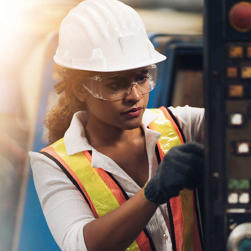 Person wearing a hard hat, safety vest, and gloves operating industrial equipment, representing workplace safety in engineering or manufacturing.