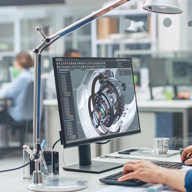 Person working at a desk with 3D mechanical design software on a computer monitor, representing digital engineering or CAD design.
