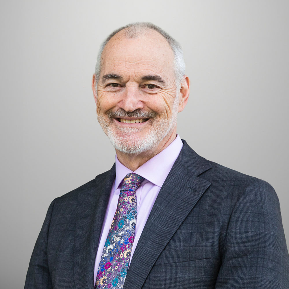 Alec is smiling towards the camera against a solid white background. Alec is wearing a light blue shirt and a necktie with a geometric pattern, underneath a navy blue blazer.