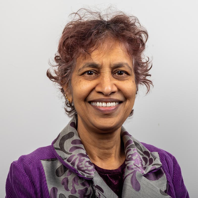 Tricia is smiling towards the camera against a solid white background. Tricia has short blonde hair and is wearing a colourful turtleneck sweater.