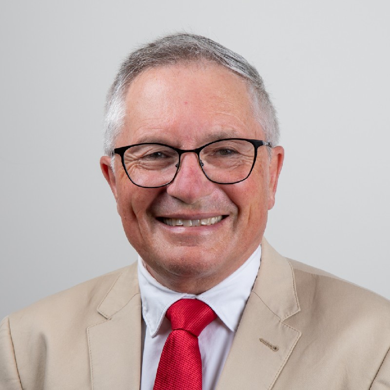 David is smiling towards the camera against a solid white background. David is wearing a white shirt and red tie, with a beige coloured blazer.