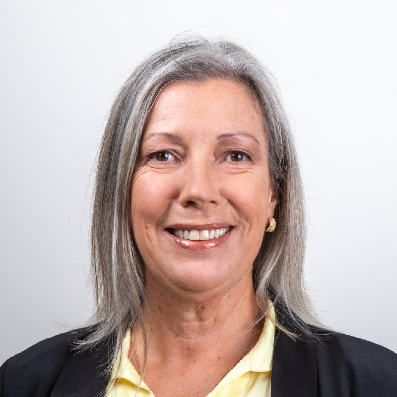 Megan is smiling towards the camera against a solid white background. Megan is wearing a yellow shirt with a black blazer.