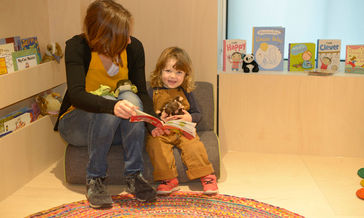An adult sits next to a child on a cushioned seat, reading a picture book together in a children’s reading area with books and toys.