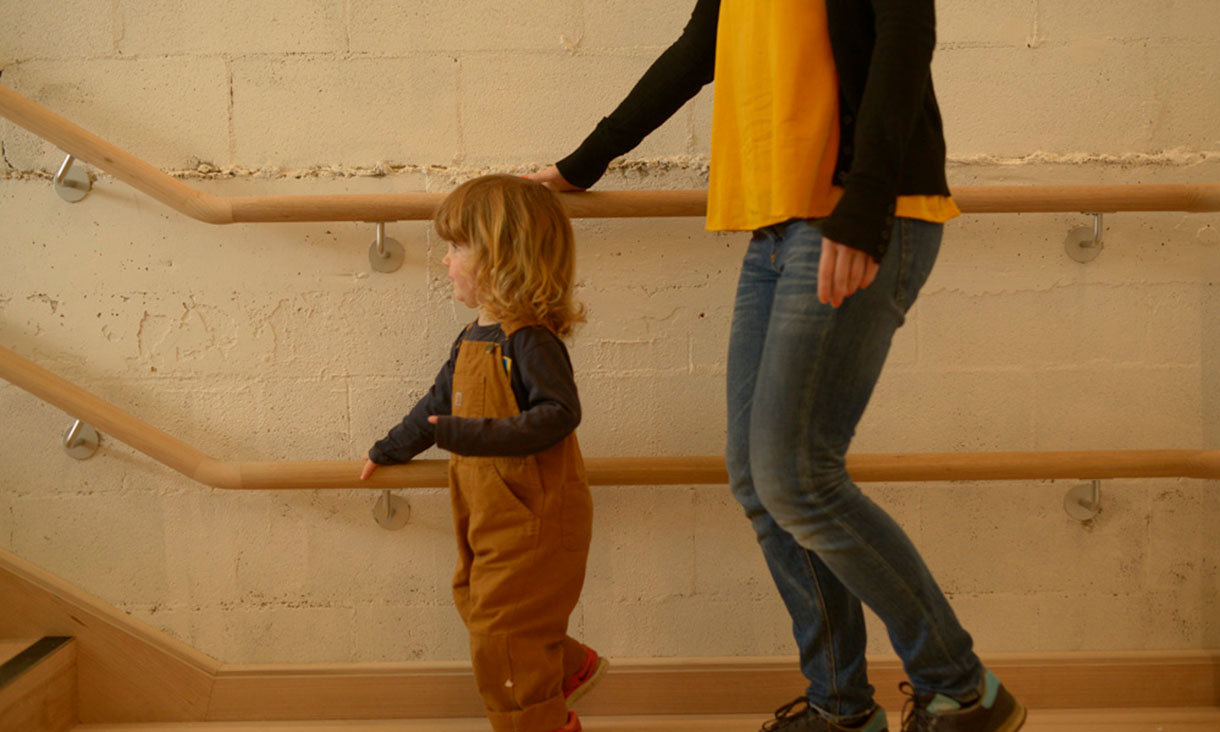 An adult and child walk up wooden stairs, holding handrails, beside a textured wall.