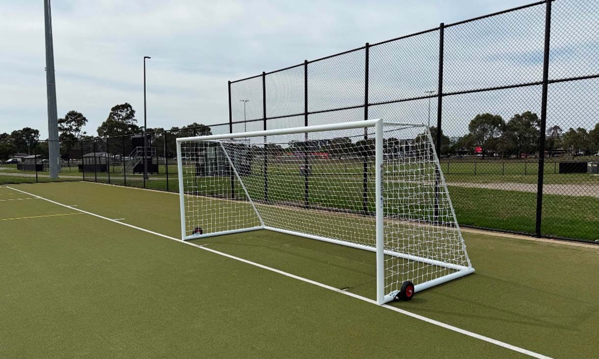 A wide outdoor synthetic turf field with soccer goals and tall perimeter fencing under a clear sky.