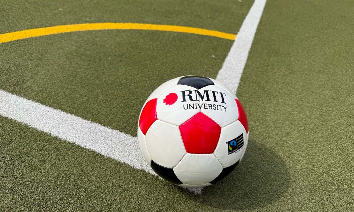 A red, white, and black RMIT-branded soccer ball resting on an artificial turf near a yellow curved field marking.
