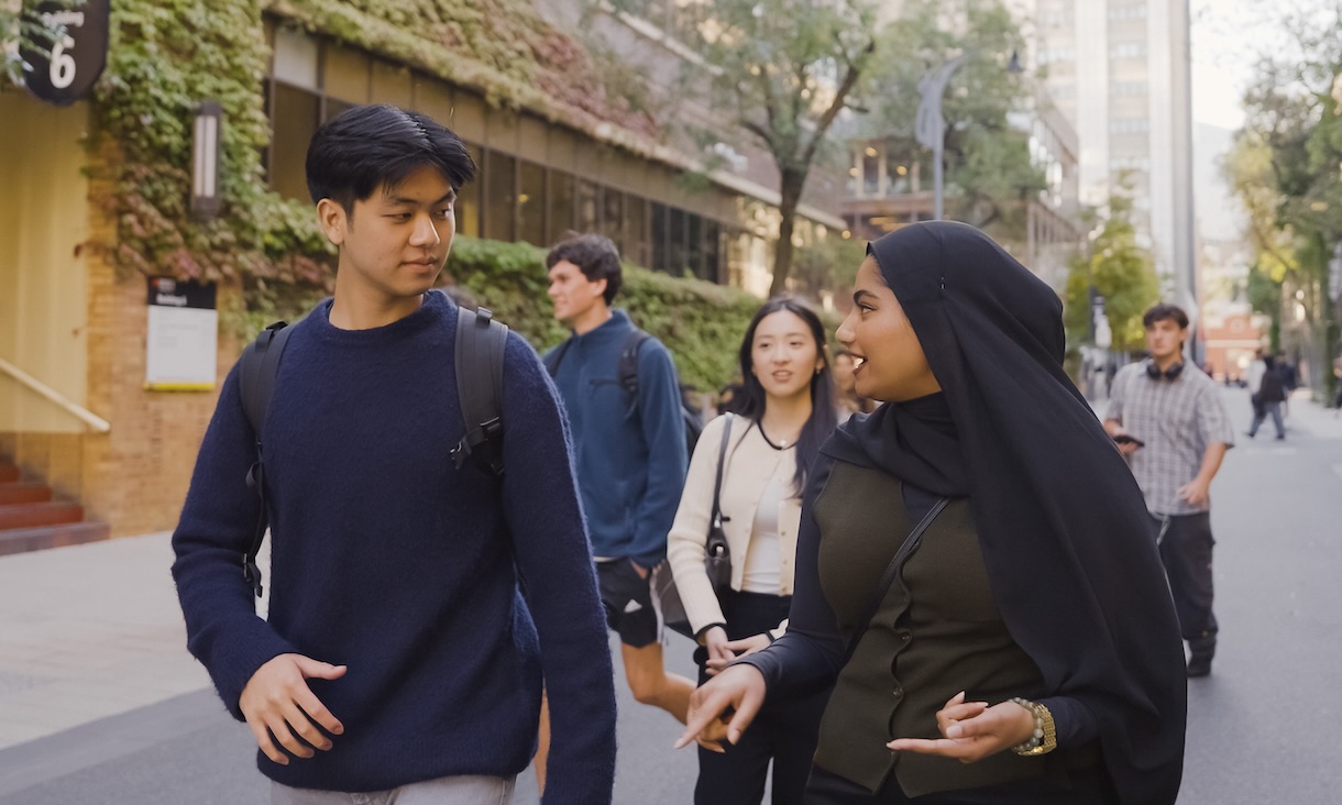 A group of students walk along an outdoor campus path, two in the foreground smiling and talking, with others walking behind them and greenery covering a building wall.