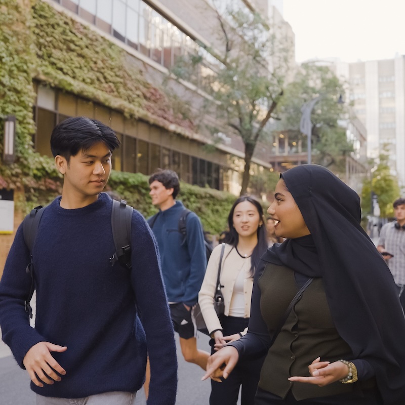 A group of students walk along an outdoor campus path, two in the foreground smiling and talking, with others walking behind them and greenery covering a building wall.