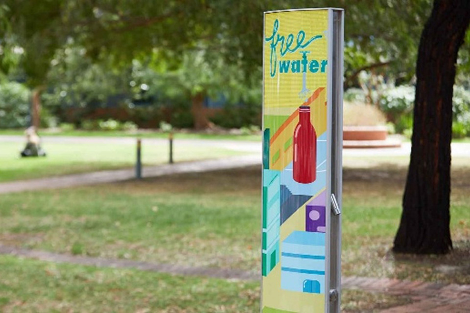 Free water refill station in a park setting, featuring a sign with a red bottle and colourful graphics inviting people to access drinking water.