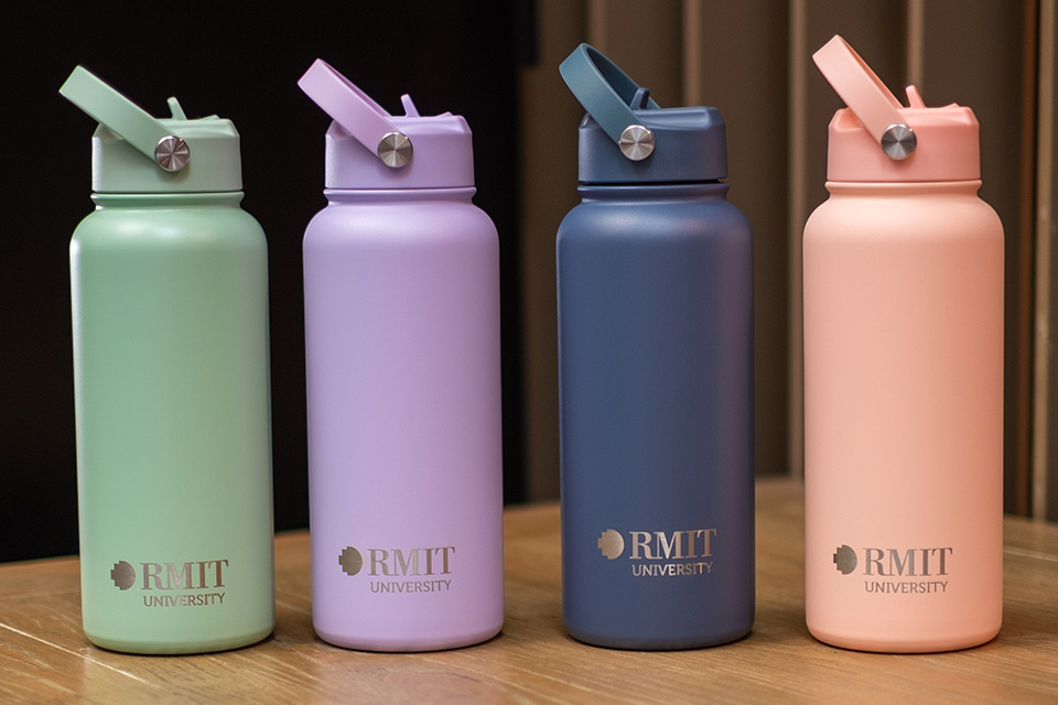 Four reusable drink bottles in pastel green, lavender, navy blue and light pink, each featuring the RMIT University logo, displayed upright on a wooden surface.