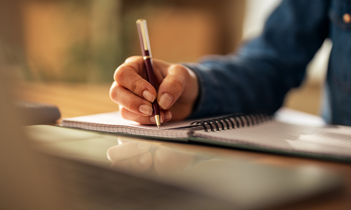 Close-up of a woman's hand as she writes in a notebook, with a pen poised above the lined pages.
