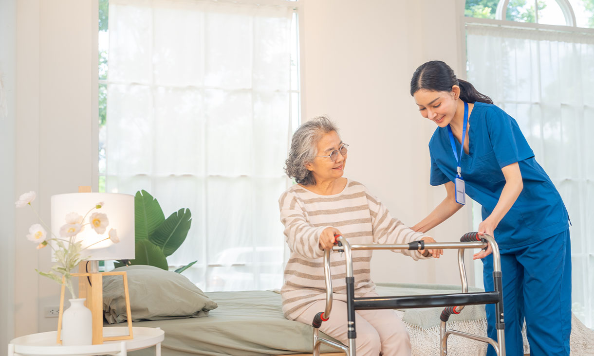An elderly woman practices walking with a wheeled walker as a smiling nurse in blue scrubs offers support in a bright care room