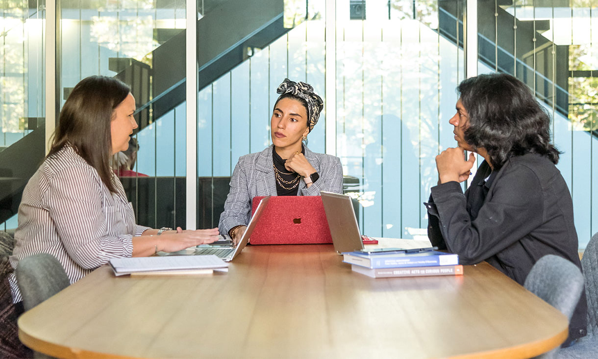 Three people sit around a table with laptops in front of them
