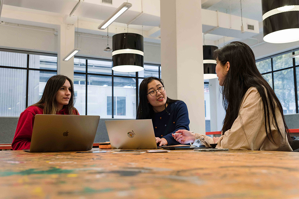 Three students sitting around a table, two have laptops in front of them