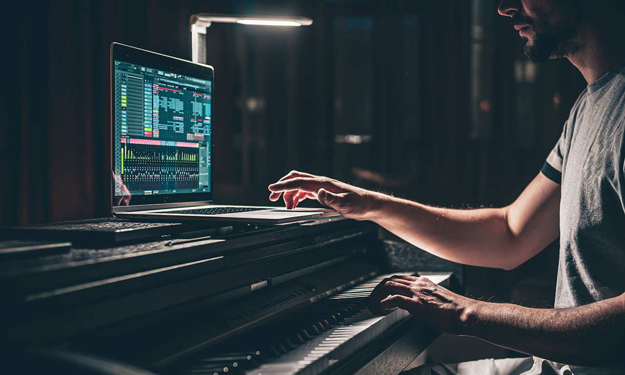 A man sits at an electric piano in a darkened room while using a laptop