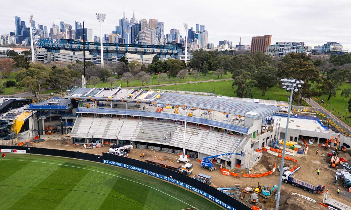 Aerial view of a sports stadium under construction beside a green park, with Melbourne’s city skyline in the background. 