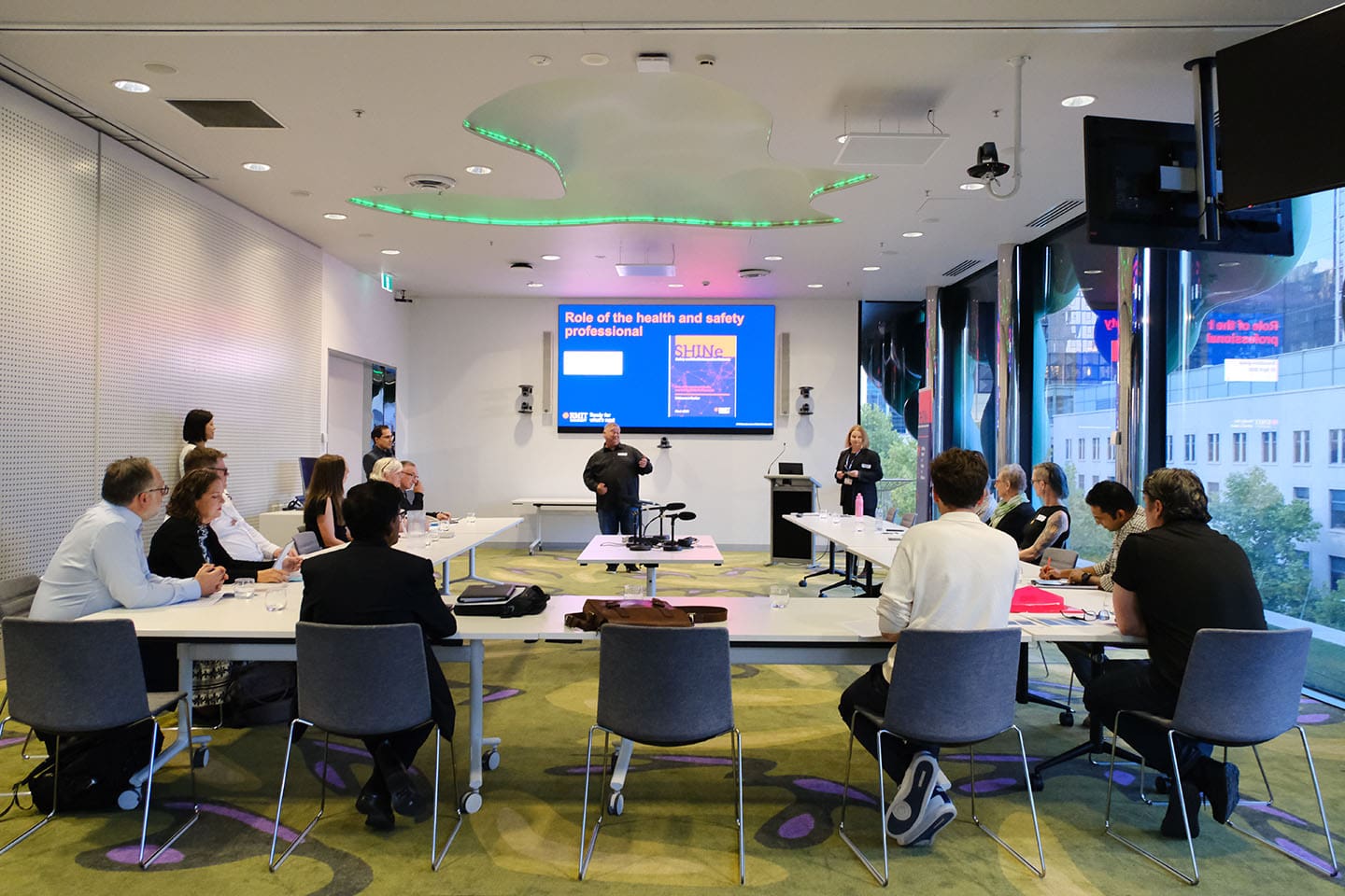 A group of people sit in a large meeting room at tables arranged in a horseshoe shape facing a screen.