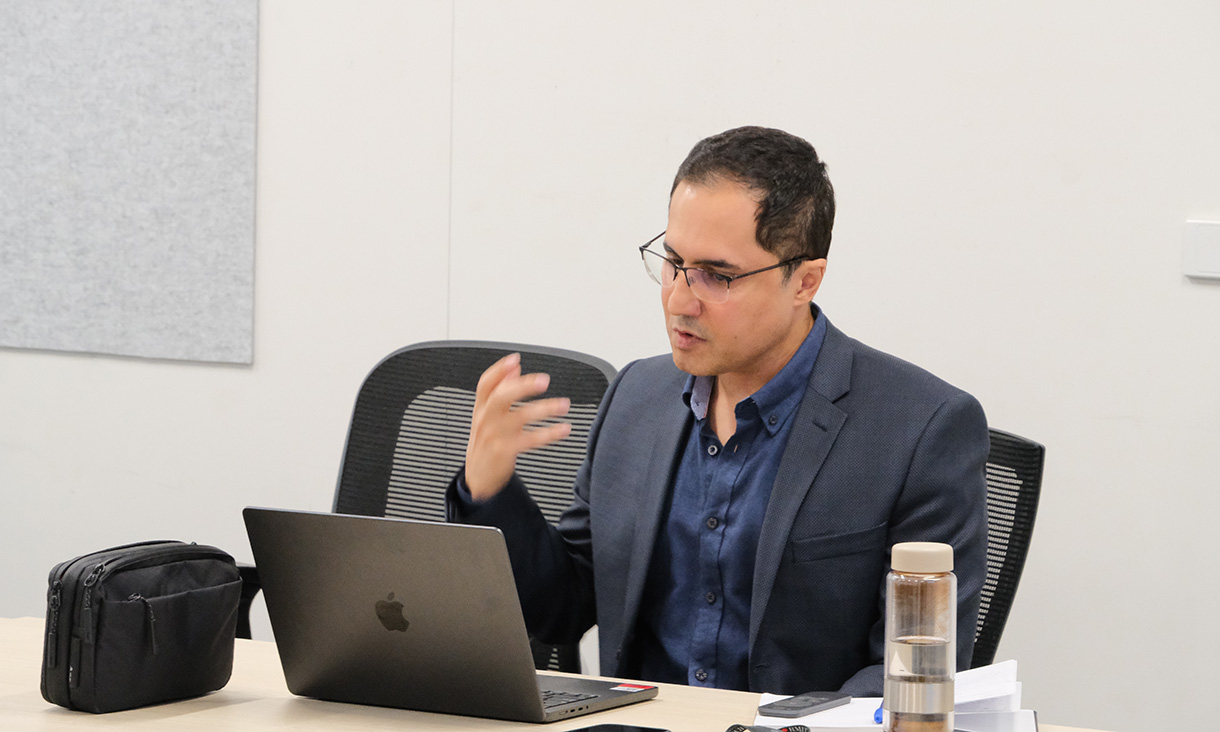 Person in a suit jacket sits at a table with a laptop, water bottle, and bag, gesturing while speaking in a modern meeting room.