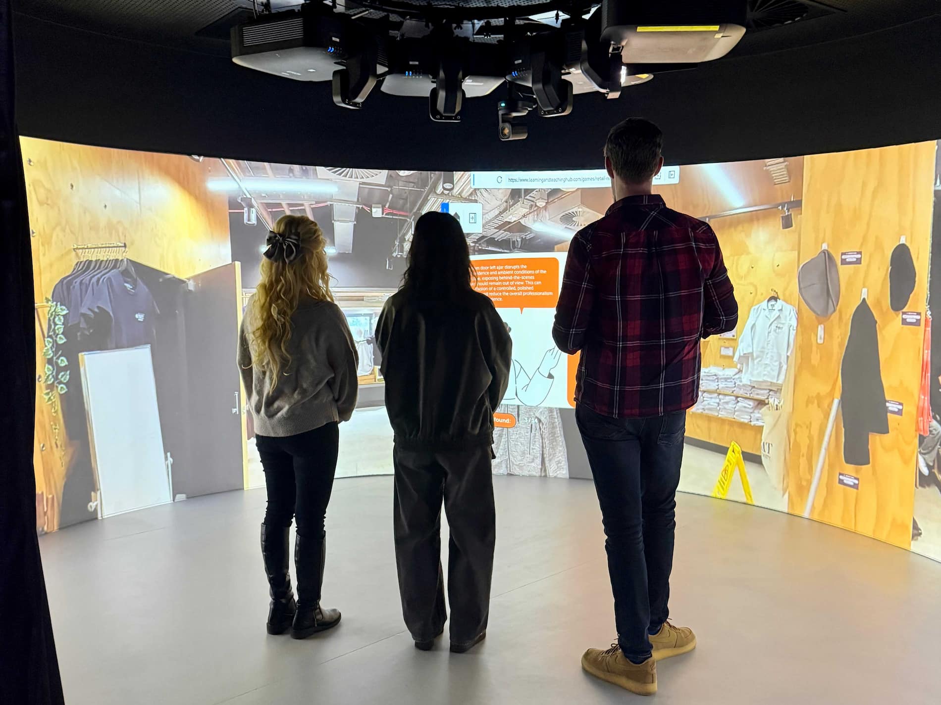 Three people stand in an immersive projection room, viewing a virtual retail-store environment displayed across curved walls.