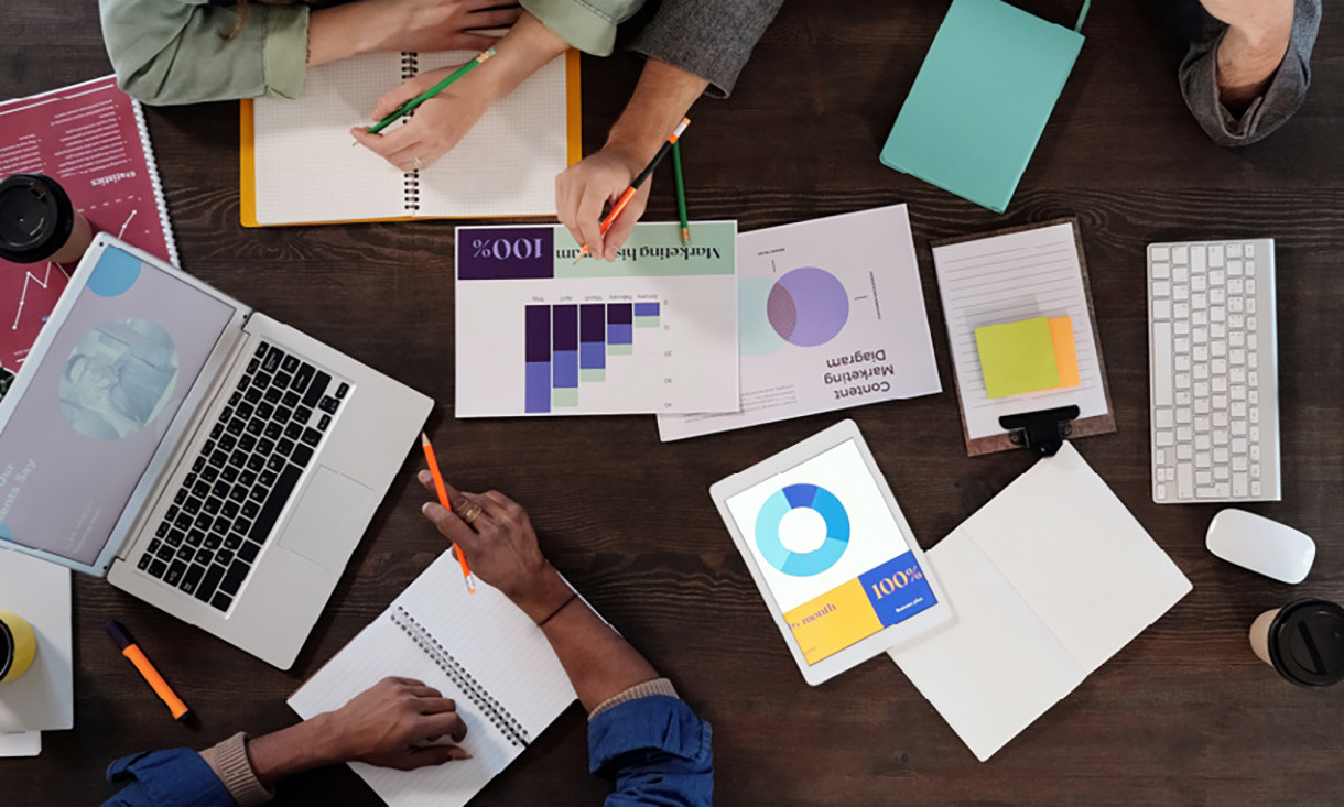People leaning across a table with laptops and papers