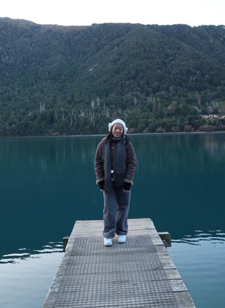 Sandra is in winter clothing and headphones and stands on a dock overlooking a calm lake and forested hills.