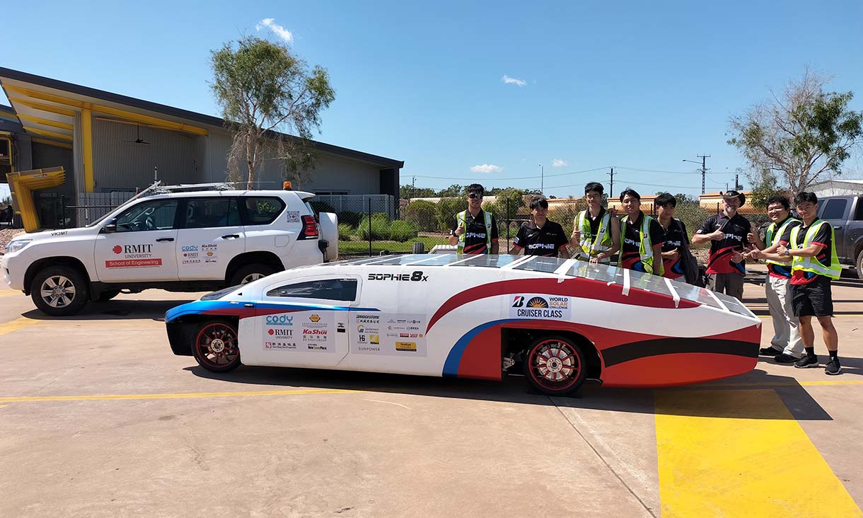 The low, aerodynamic vehicle is covered in solar panels and sponsor logos, including RMIT University, with a white RMIT School of Engineering support SUV parked behind it and an industrial building and trees in the background.