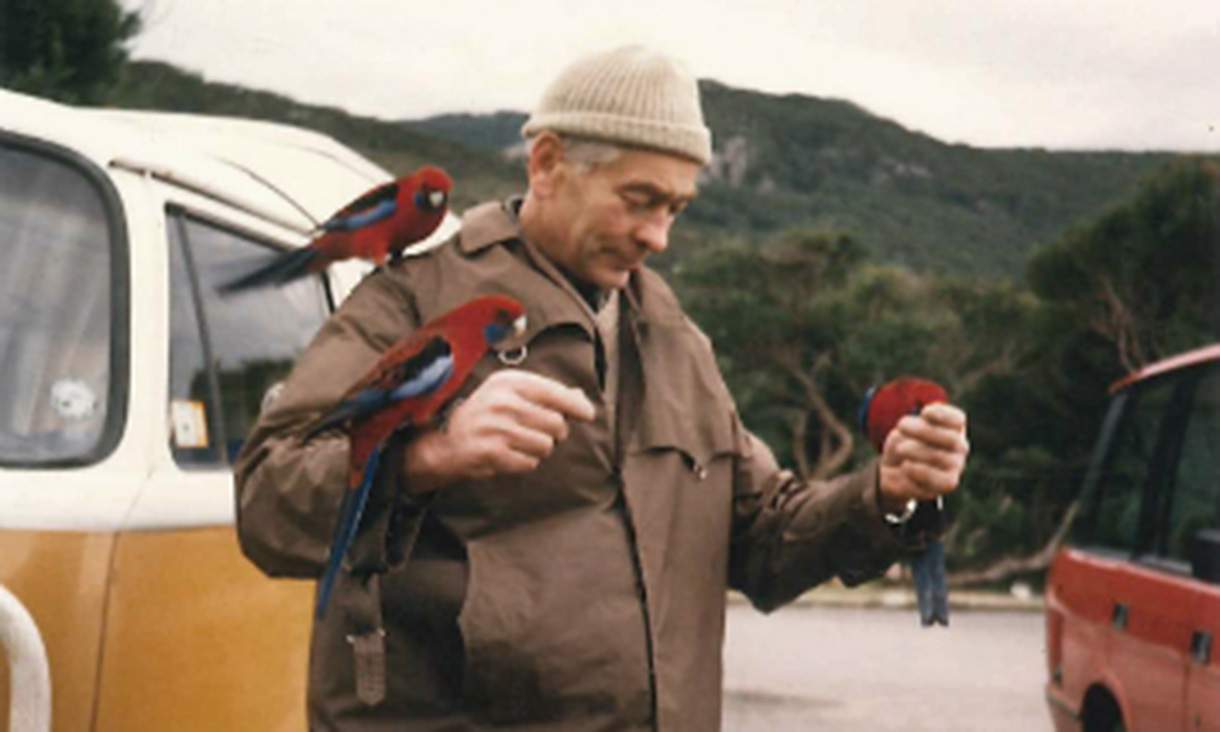 A person in a brown jacket and knitted beanie stands outdoors near vehicles, holding and feeding bright red and blue parrots perched on their hand and shoulder, bushy hills in the background.