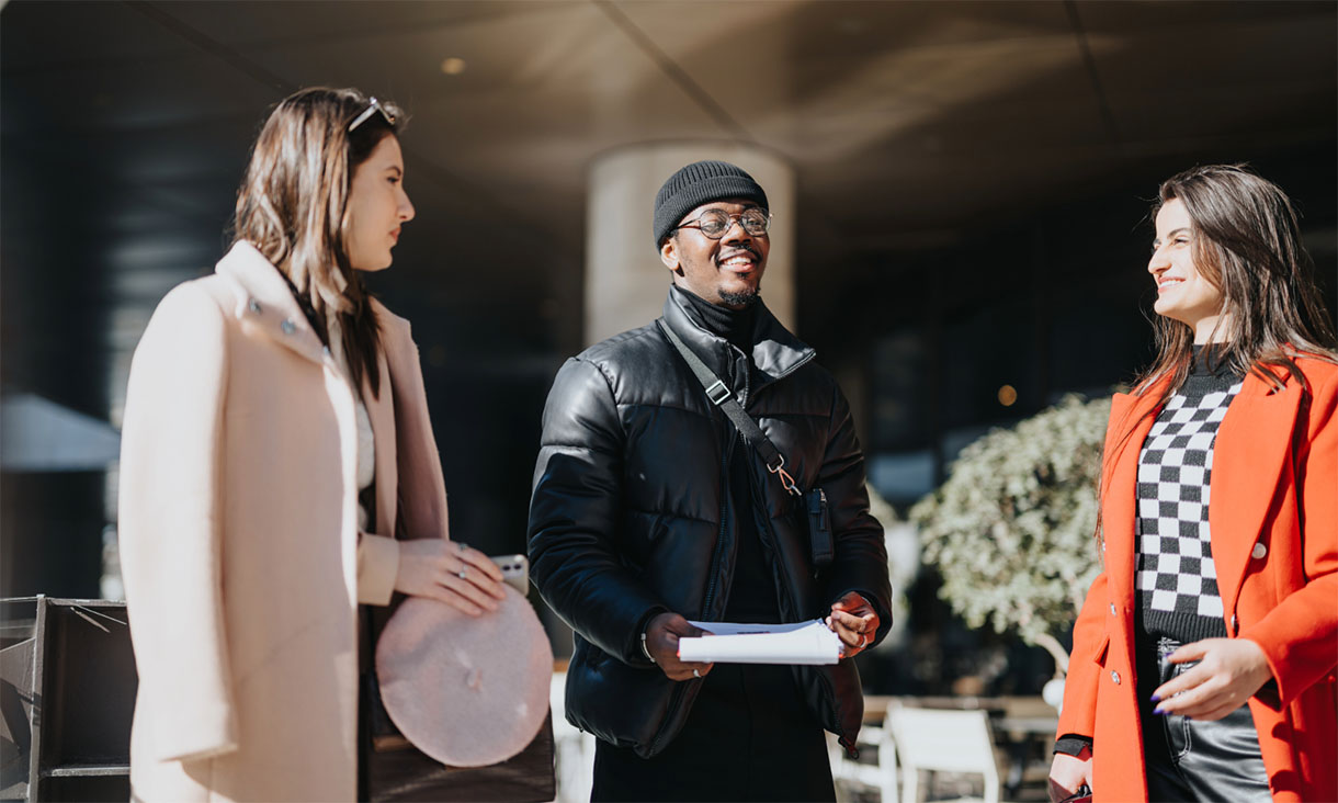 Three people with blurred faces stand talking outside an urban building; the person in the centre wears a black puffer jacket and holds papers, while the two others wear light and bright-coloured coats.