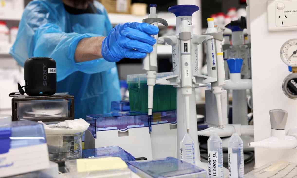Photograph of various laboratory materials and tools on a desk. In the middle of the photograph a man wearing protective, blue clothing and gloves is reaching for one of the tools. 