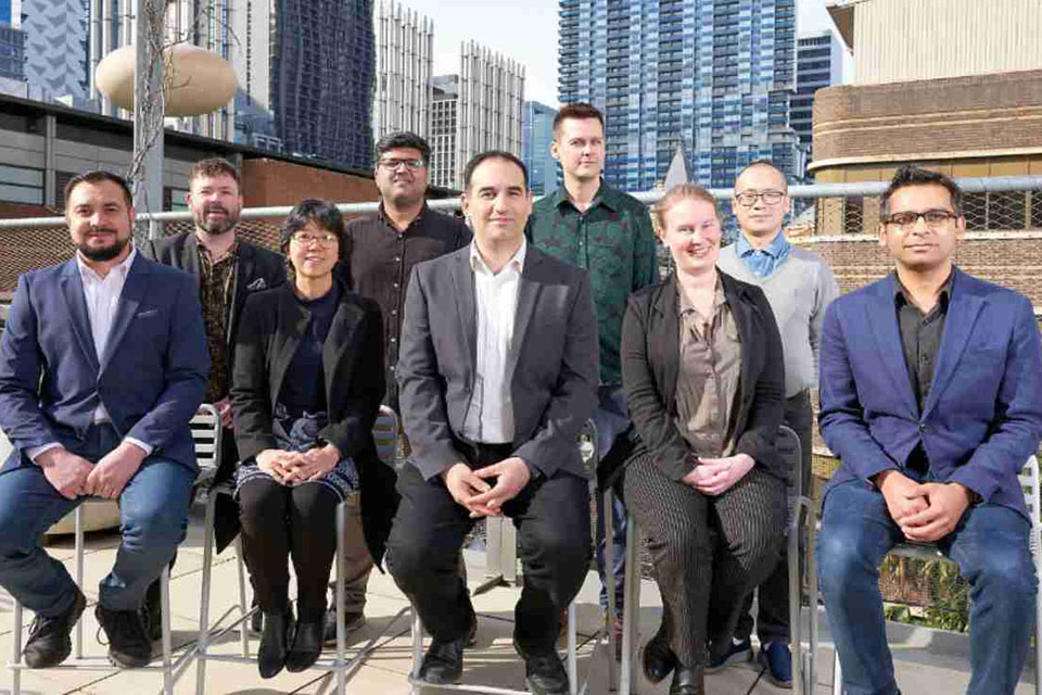 Photograph of the comas team. There are people people featured. Five sit at the front and four stand in the back. In the background there are various tall buildings.