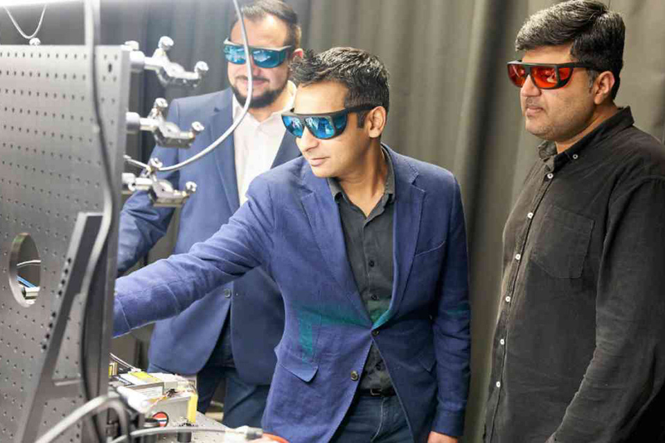 Photograph of three men standing in a workshop. The man in the middle is fiddling with a piece of equipment. All three men wear protective glasses. 