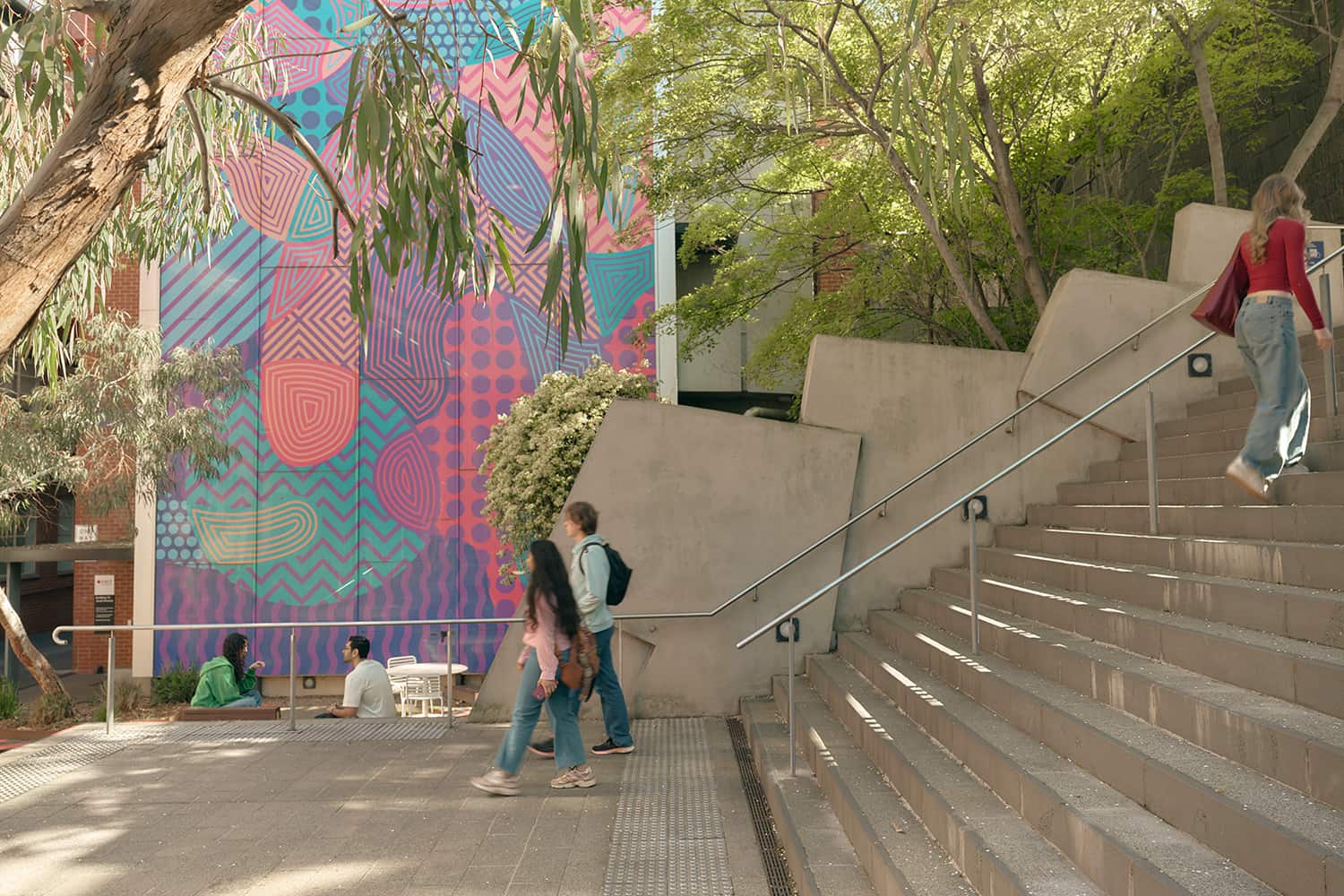 Photograph of Ngarara Place at RMIT city campus, an outdoor urban space featuring a colourful geometric mural with patterns in pink, blue, and purple on the side of a building. Several people are walking or sitting near concrete stairs surrounded by trees and greenery in dappled sunshine.