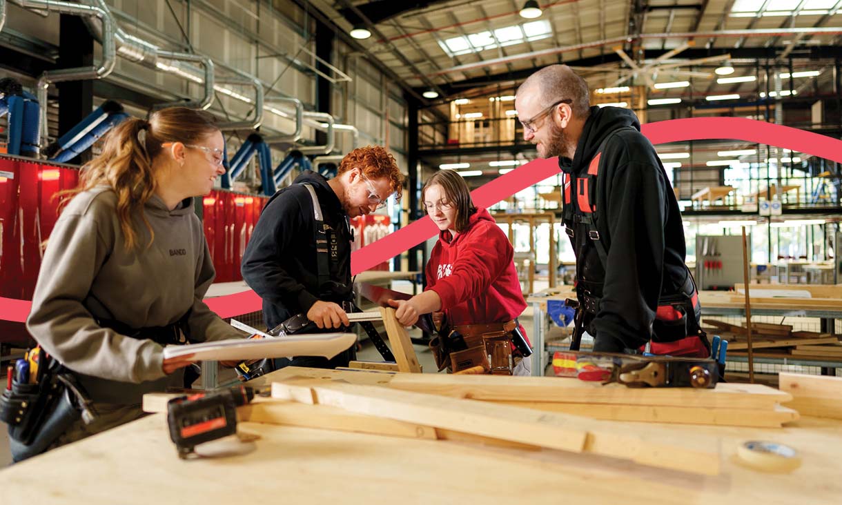 Group of people in a workshop wearing tool belts and safety gear, working together with timber and tools on a large workbench in an industrial training environment.