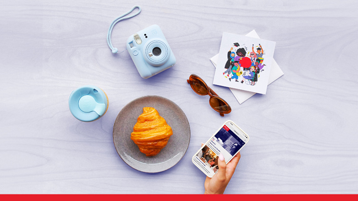 Flat lay of a croissant on a plate, a hand holding a smartphone, and nearby items including a blue instant camera, a reusable cup, sunglasses and a colourful card on a light wooden background.