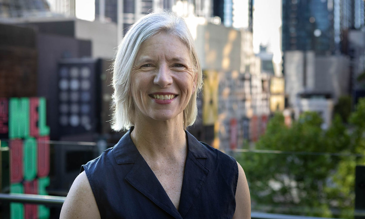Dianne standing outdoors wearing a sleeveless top, with a city skyline and trees visible in the background.