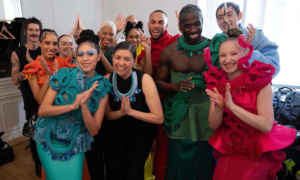 Group of people posing together in vibrant, sculptural outfits made from textured fabric in red, green, blue and orange, standing in a well-lit room and making playful hand gestures.
