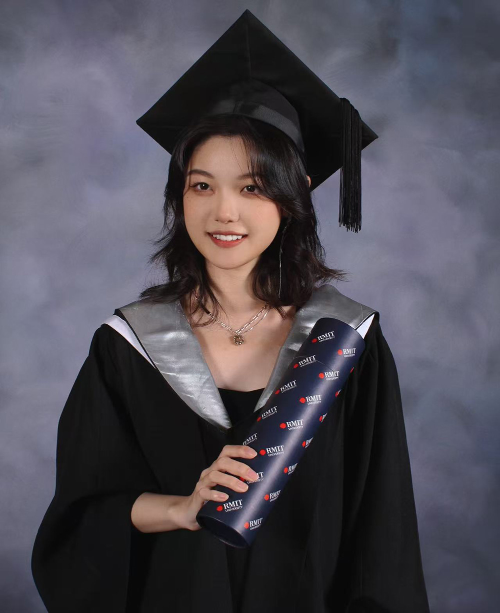Rita in her graduation cap and gown holding a rolled RMIT University certificate folder, posed in front of a grey studio backdrop
