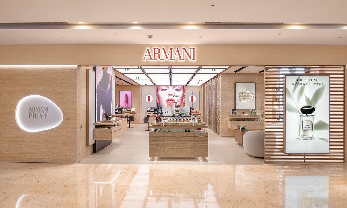 Front view of an Armani beauty store with light wood interior, illuminated ‘ARMANI’ sign above the entrance, central display counters and large perfume and cosmetic posters on the walls.