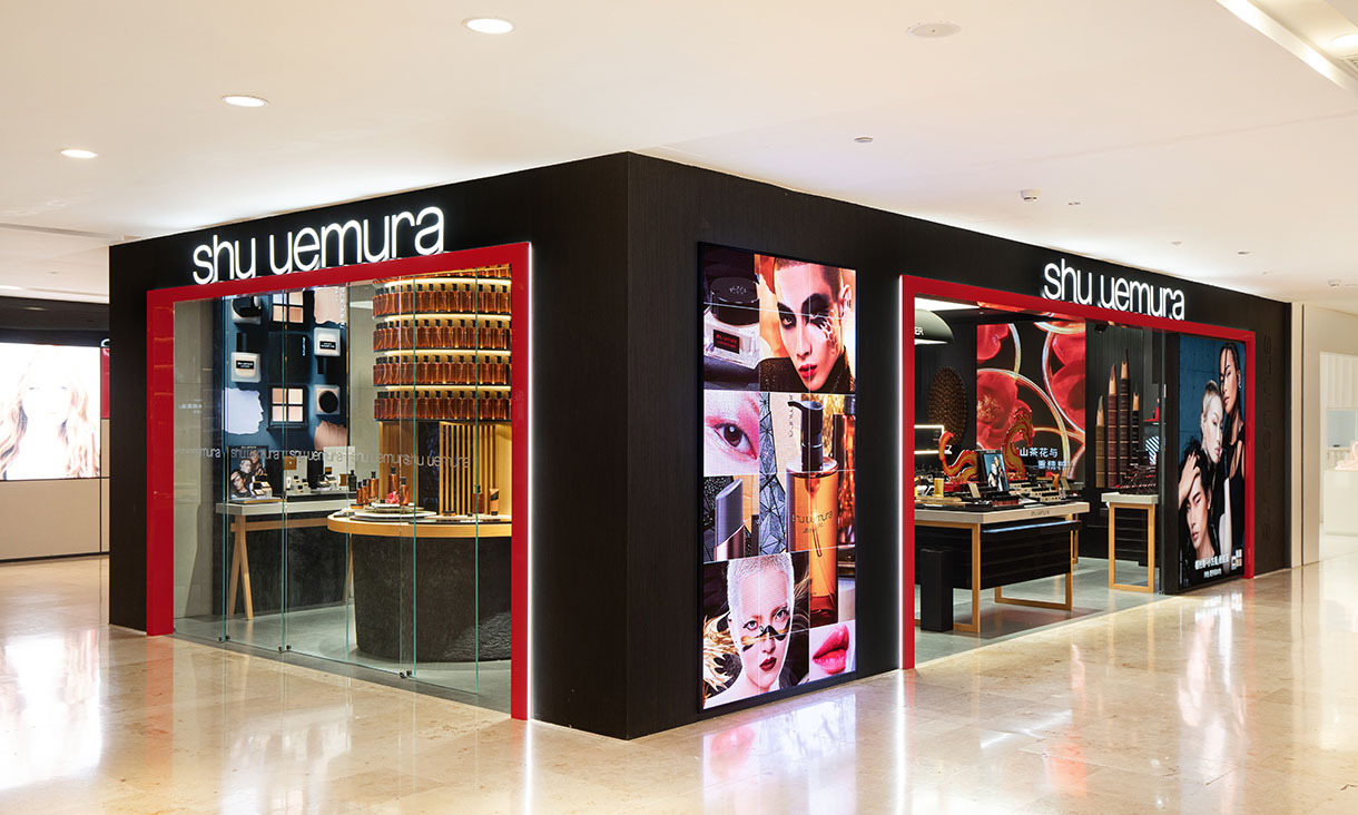 Corner view of a Shu Uemura cosmetics store with black exterior framing, red accents, glass display windows and large illuminated images of makeup products and faces.