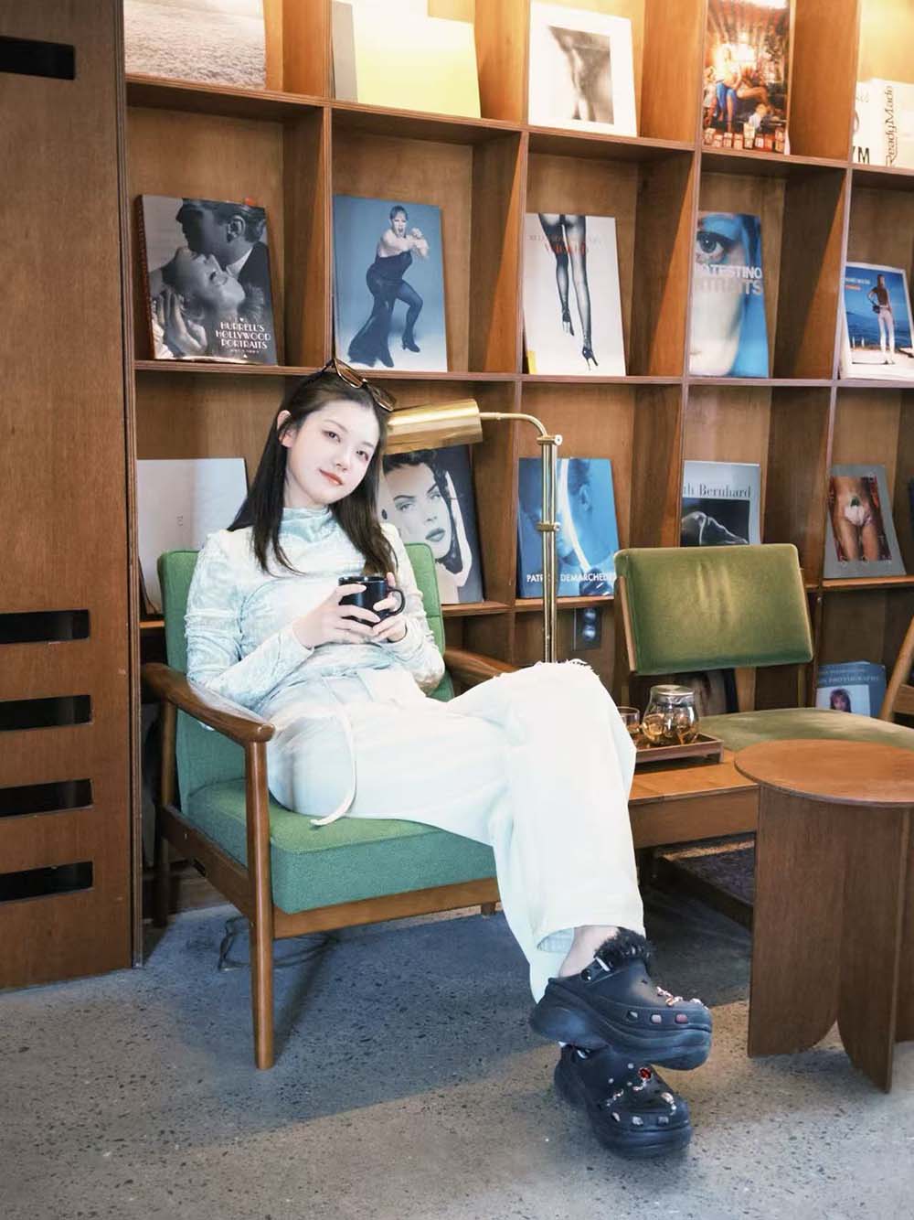 Rita is sitting in a green armchair. She is holding a mug with her legs crossed and is surrounded by wooden shelves filled with large photography and fashion books.