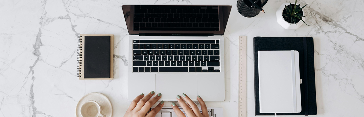Hands over a laptop, with a notebook and pens next to them
