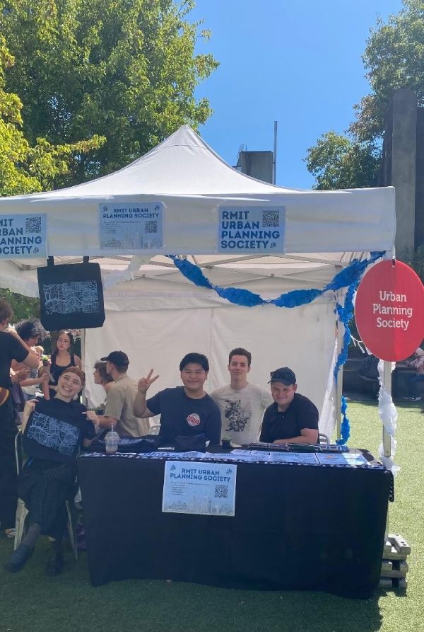 Bethany, Jorge, Tristan and Tom are sitting at a table under a white marquee. There are RMIT Urban Planning Society signs stuck on the front. They are all smiling.
